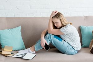 depressed teen girl sitting on couch with smartphone while doing homework